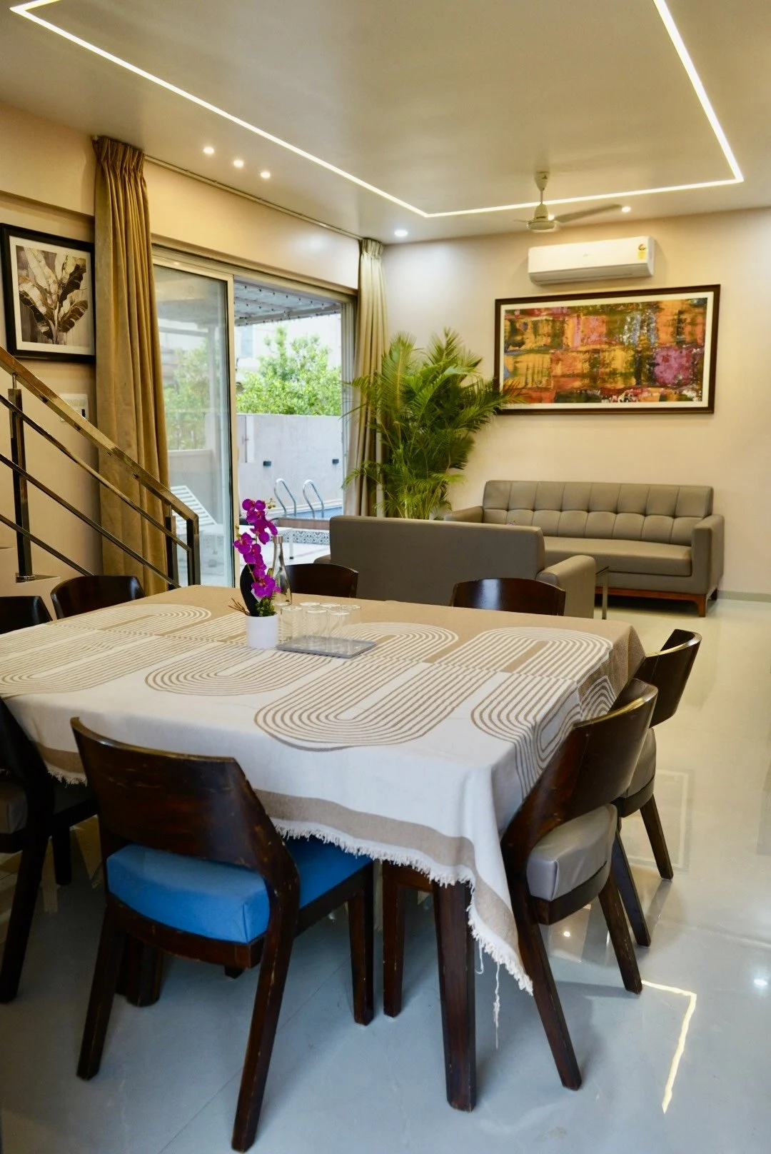 A dining table with a patterned cloth, chairs and purple flowers, beside the lounge and sliding doors overlooking the pool.