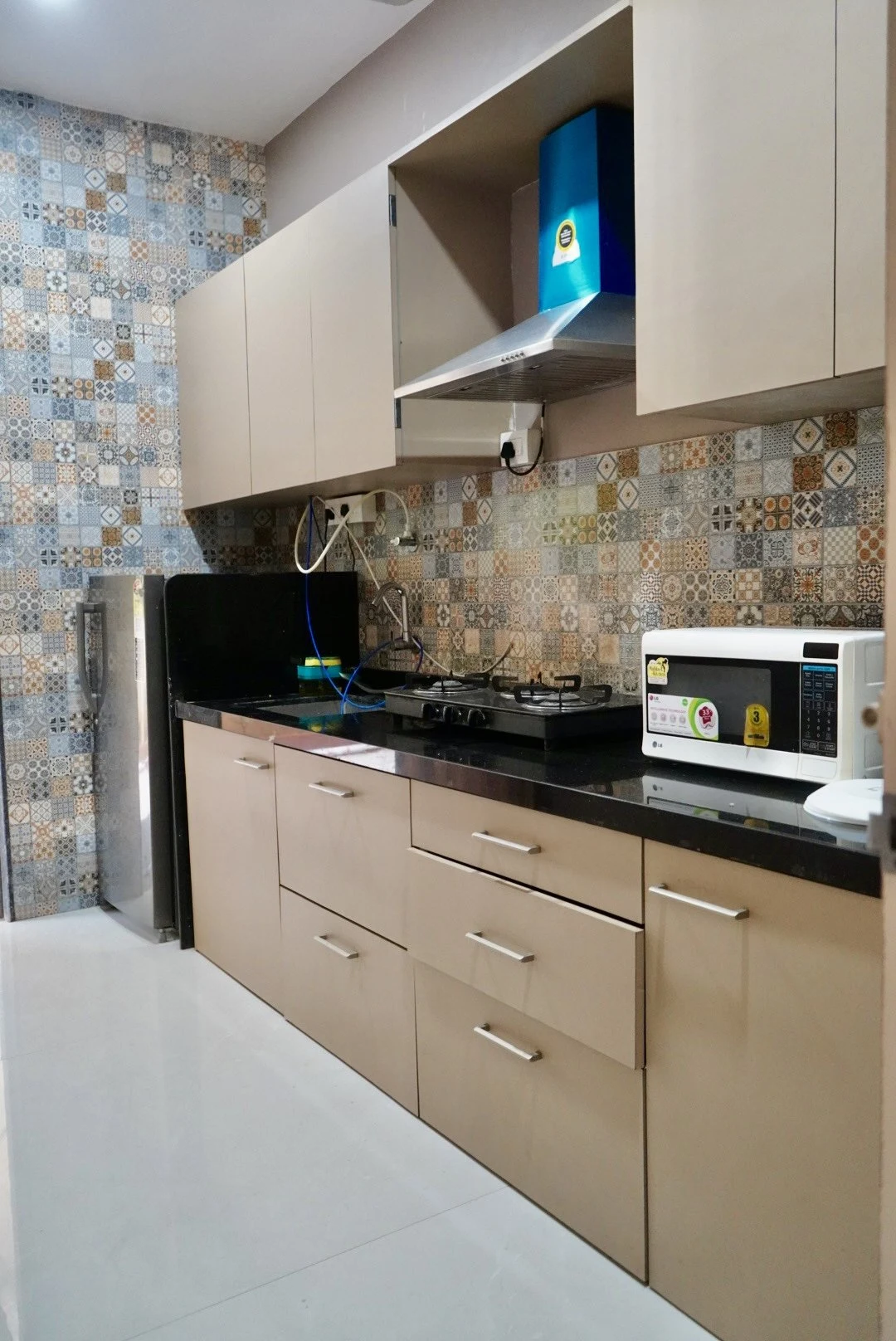 A kitchen with pale cabinets, patterned tiles, a black countertop, gas hob, sink, microwave and refrigerator.
