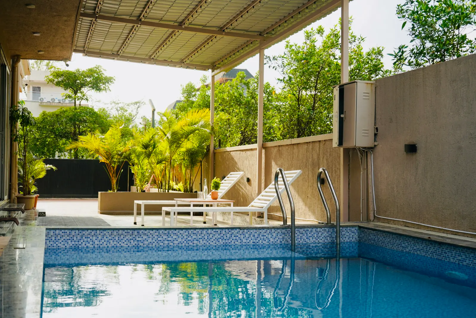 Aashiyana’s blue-tiled pool in daylight, with a metal ladder, white loungers, a covered deck and green plants.