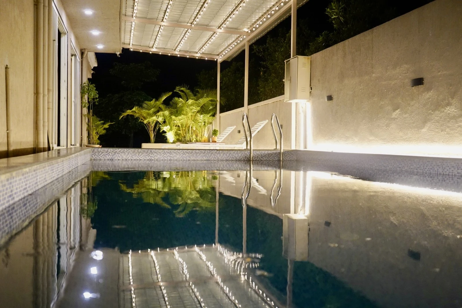 The private pool at night, reflecting string lights from the covered deck, with palms and a metal ladder beyond.